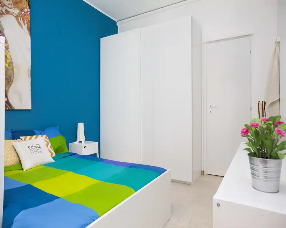 Bright, modern bedroom with a colorful bedspread, white wardrobe and minimalist furnishings creating a fresh, welcoming atmosphere.