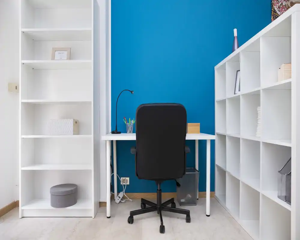 Neat home office corner with a white desk, ergonomic black chair and modular shelving against a vivid blue accent wall — functional, minimal and well organized for focused work.