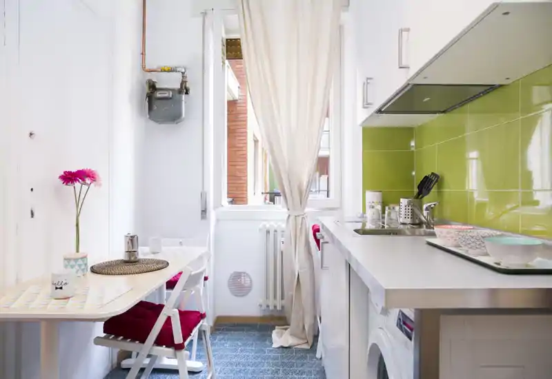 Bright compact kitchen with a small dining area: white countertops, green tiled backsplash, sink and washing machine, plus a window that brings natural light. Perfect for a cozy city apartment with practical layout and colorful accents.