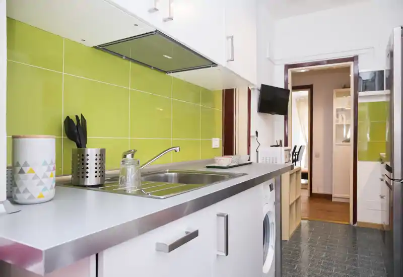 Compact modern kitchen with green tiled splashback, stainless sink, and integrated appliances—bright and neatly arranged for short-term stays.