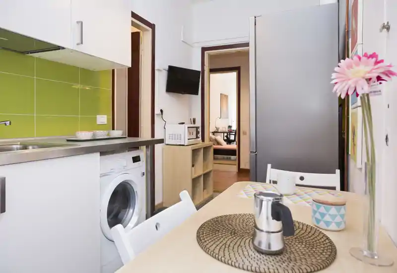 Bright, compact kitchen with green tiled backsplash, countertop sink, washing machine and a small dining table neatly set with a coffee maker and cups.