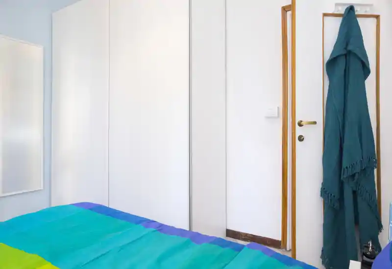 Bright, compact bedroom with a colorful bedspread and large white wardrobe; clean minimalist styling and a hanging teal throw on the door.