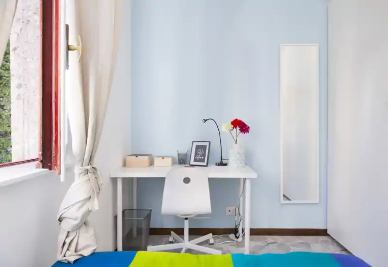 Small, bright bedroom corner with a white desk and chair facing a window; neat decorative touches include a vase with flowers and a framed print. The bed is only partially visible in the foreground, and the space feels tidy and functional for work and rest.