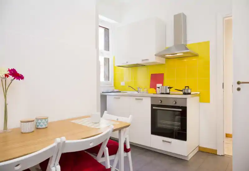 Compact modern kitchen with a bright yellow tile backsplash, white cabinets, built-in oven and a small wooden dining table — stylish and well lit, ideal for a cozy city apartment.