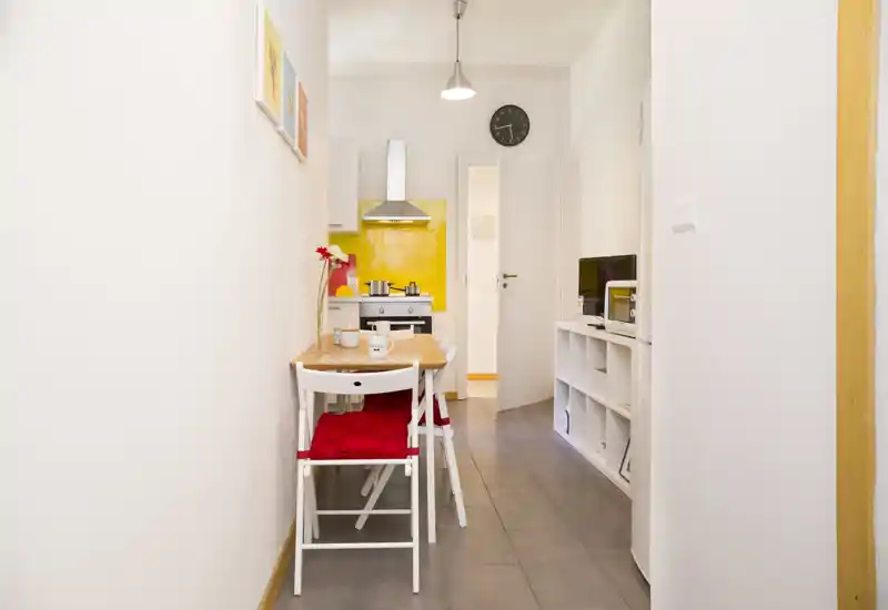 Compact fitted kitchen with a bright yellow backsplash, small dining table and two chairs; neat, well-lit space ideal for a studio apartment.