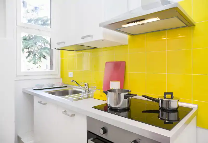 Modern compact kitchen with a bright yellow tiled backsplash, induction hob with stainless pots, stainless sink, and white cabinets. Natural light from the window and clean, contemporary finishes make this kitchen feel fresh and functional.