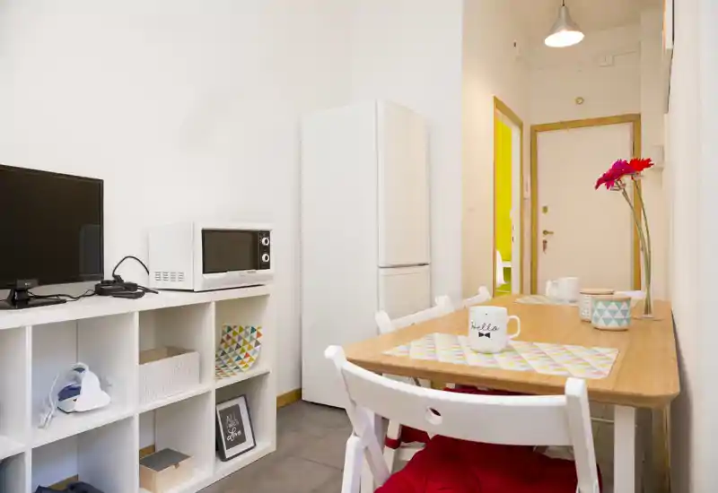 Bright, compact dining area with a wooden table set for two, decorative placemats and fresh flowers; kitchenette shelving and appliances visible, ideal for a small apartment.