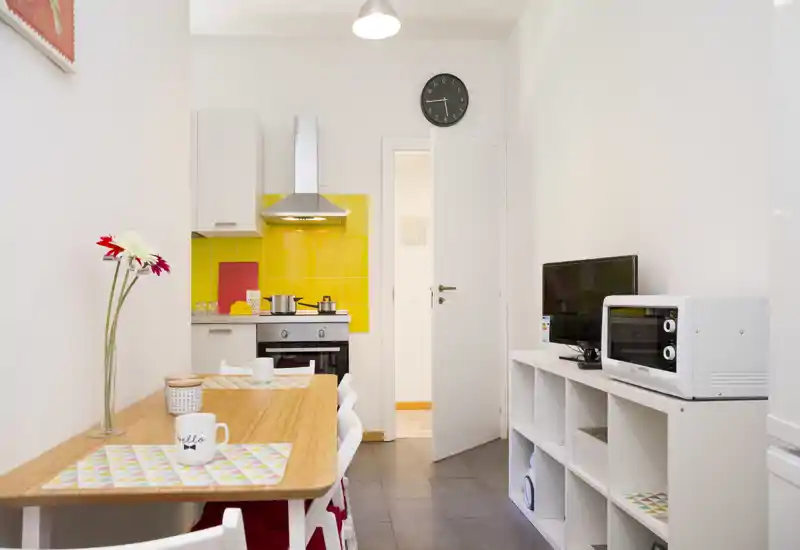Bright, compact kitchen with a wooden dining table, colorful placemats and yellow tile backsplash. Includes oven, hood, microwave and a small TV — ideal for a cozy city apartment.