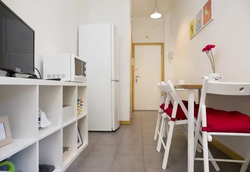Compact kitchenette with a small dining area: white cabinets, fridge and microwave on the left and a narrow table with red-cushioned folding chairs on the right. Bright, tidy and efficiently arranged for short stays.