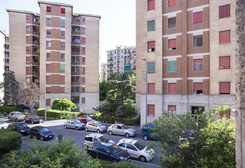 Vista luminosa de la calle y edificios residenciales con coches aparcados, vegetación y fachadas — ideal para mostrar la ubicación y el contexto del barrio.