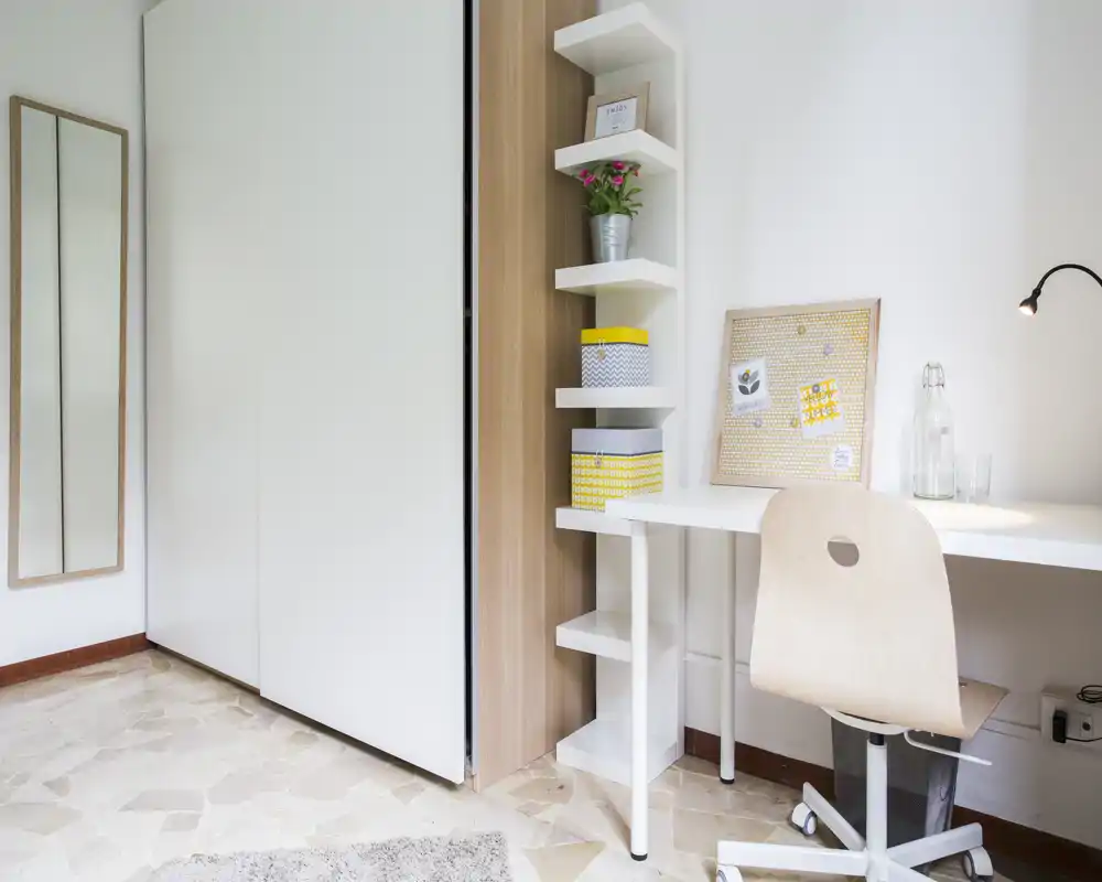 Bright, minimal bedroom corner with a white wardrobe, built-in shelving and a tidy desk with a wooden chair — ideal for focused work or study.