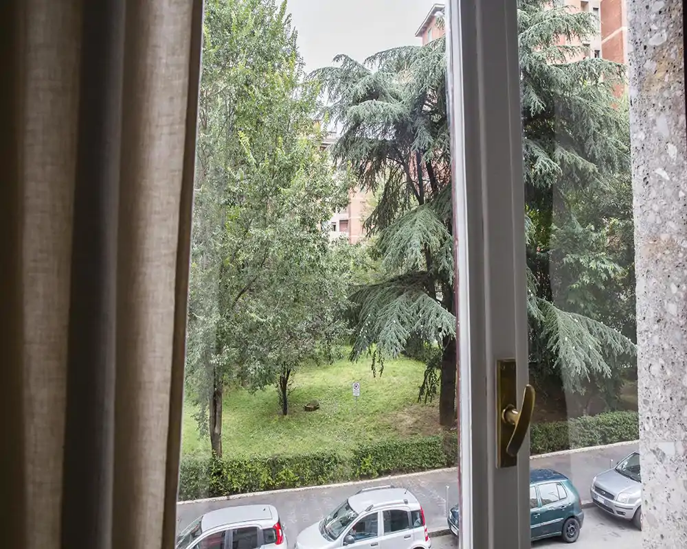 Window view overlooking a small green courtyard with mature trees and parked cars along the street — bright, refreshing outdoor aspect visible from the room.