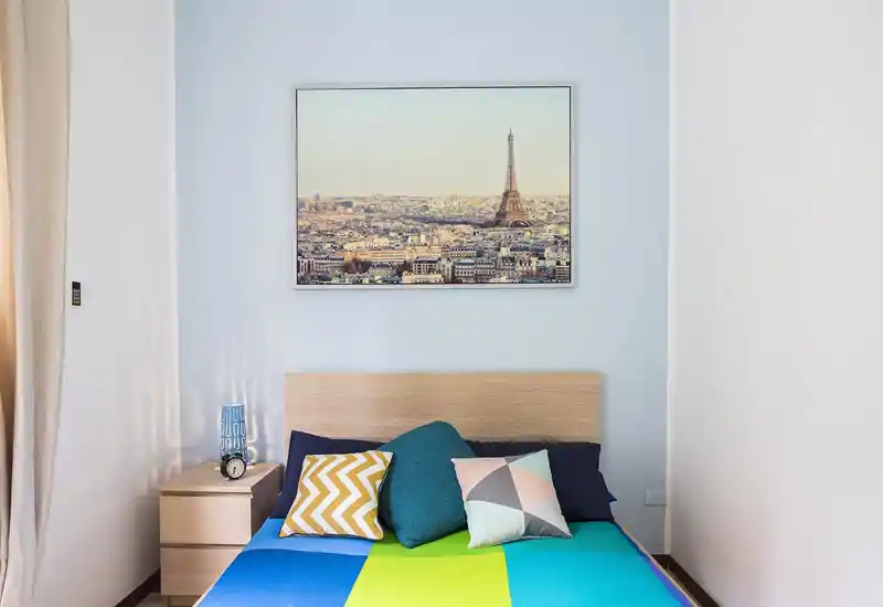Bright, neatly staged bedroom with a wooden headboard, colorful bedding and decorative cushions; framed city artwork above the bed and a bedside table with lamp create a welcoming atmosphere.