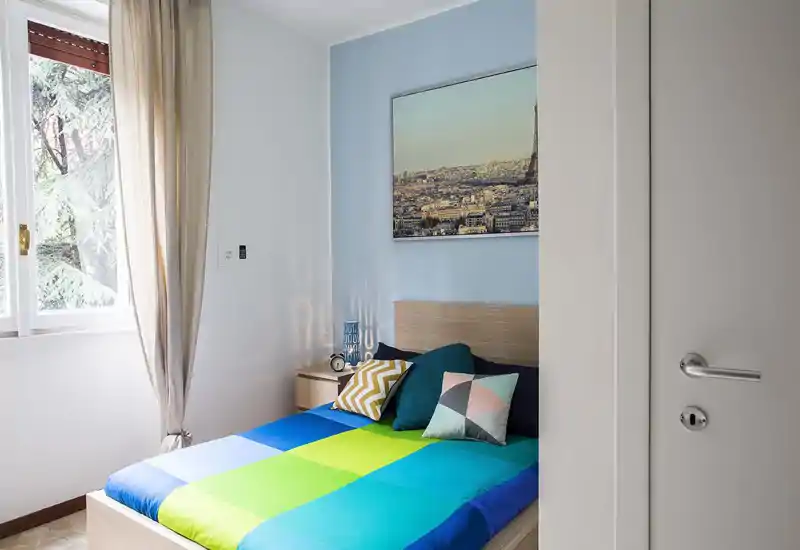 Bright, compact bedroom with a colorful double bed, decorative cushions and a bedside table; natural light from a large window creates a fresh, welcoming atmosphere.