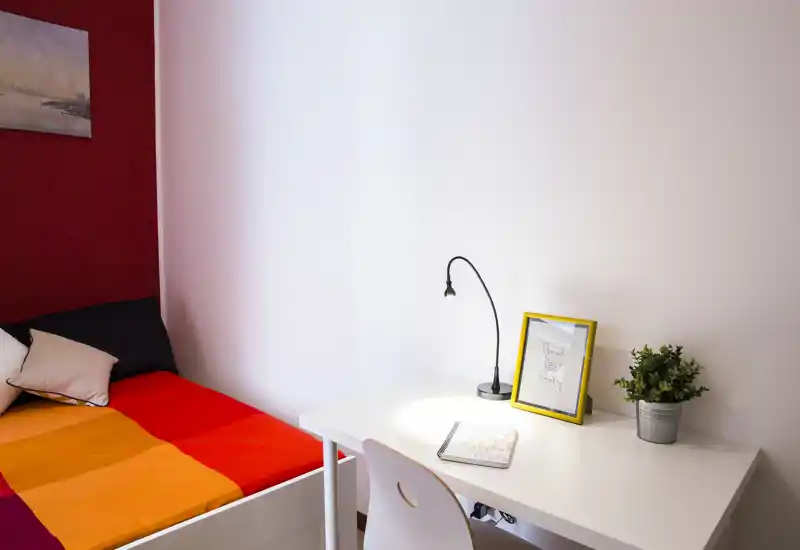 Camera da letto luminosa e compatta con un letto dai colori vivaci e una scrivania bianca ordinata. L'angolo studio presenta una lampada, una cornice e una piccola pianta, ideale per studenti o professionisti.