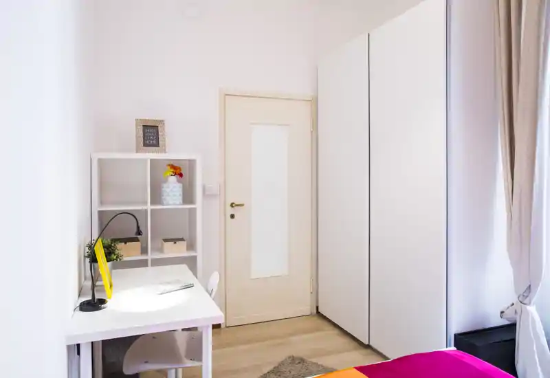 Angolo di camera luminoso e minimal con una scrivania, scaffale e vista parziale del letto. Spazio in tonalità neutre, luce naturale e arredo moderno e ordinato.