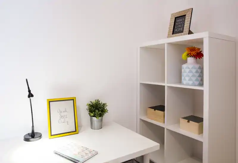 Coin bureau lumineux et minimal avec table blanche, lampe de travail, cadre et étagère avec boîtes décoratives et vase — idéal pour un petit bureau à domicile ou un coin étude.