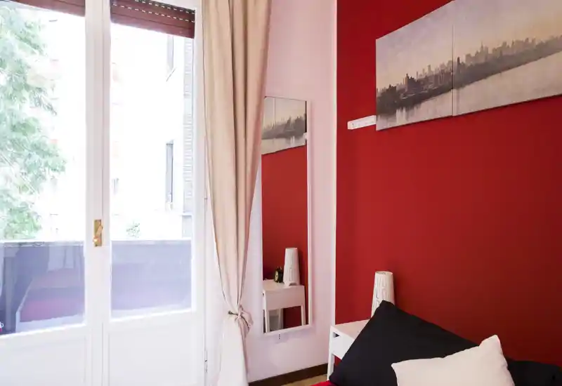 Angolo di camera luminoso e accogliente con parete rossa a contrasto, quadro decorativo e porta finestra che dà su un piccolo balcone. Il letto con cuscini scuri e chiari è visibile in primo piano, con comodino e specchio.
