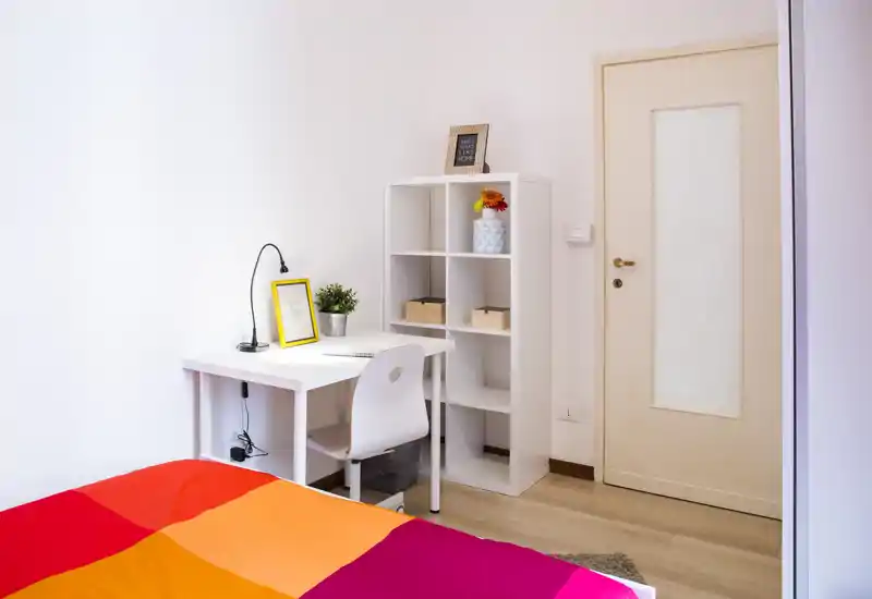 Angolo camera luminoso e ordinato con una coperta colorata, una scrivania bianca con sedia e una libreria a cubi con scatole decorative e una pianta. Lo spazio è ben illuminato e accogliente, ideale per un monolocale.