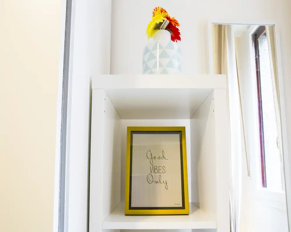 Primo piano di una nicchia a muro bianca con una cornice che riporta “Good Vibes Only” e un piccolo vaso decorativo con fiori; a destra si vede il bordo di una finestra. L'immagine mette in risalto dettagli d'arredo anziché una stanza completa.