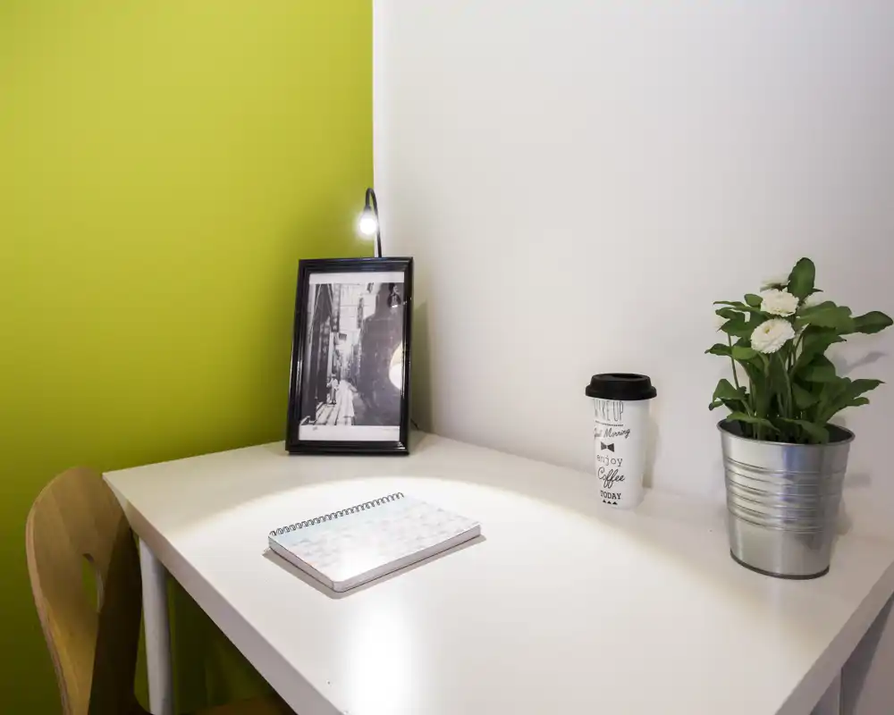 Angolo scrivania luminoso e minimal con taccuino, foto incorniciata, bicchiere da asporto e vaso con pianta — uno spazio di lavoro ordinato perfetto per soggiorni brevi.