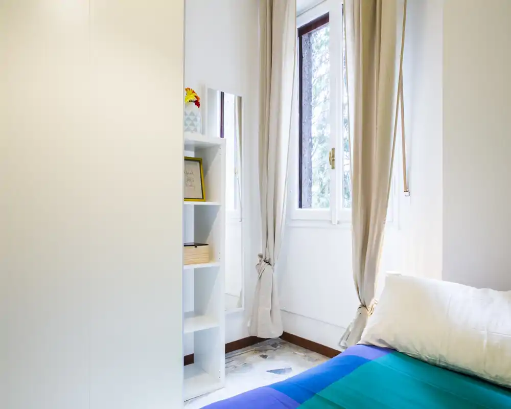 Angolo di camera luminoso e semplice con letto ben rifatto, tende morbide e luce naturale dalla finestra; scaffalatura compatta accanto al letto per riporre oggetti.