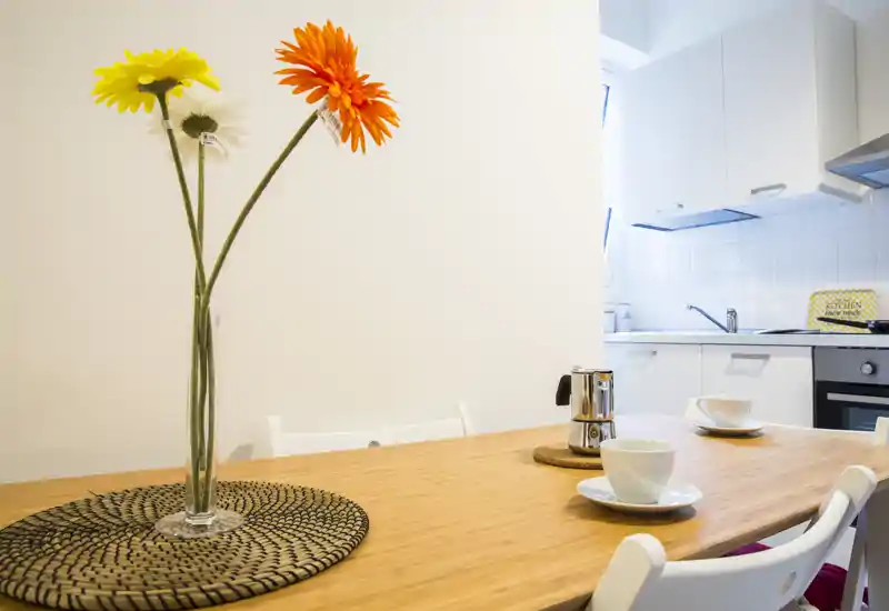 Zona pranzo luminosa e curata adiacente a una cucina compatta bianca. Un tavolo in legno apparecchiato con tazze e un vaso decorativo crea un angolo accogliente per la colazione.