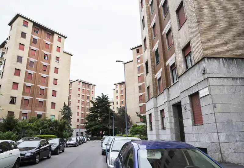 Veduta urbana a livello strada con palazzi residenziali, auto parcheggiate e una strada alberata — utile per mostrare l'esterno e il quartiere di un immobile in affitto.