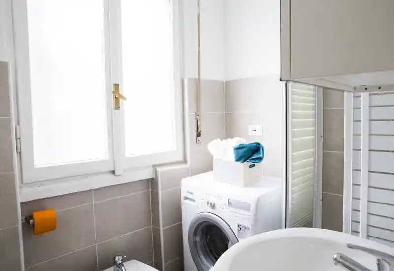Bagno compatto e luminoso con lavatrice, pareti piastrellate e un lavabo in primo piano. La luce naturale da una grande finestra e lo spazio ordinato lo rendono funzionale per un affitto.