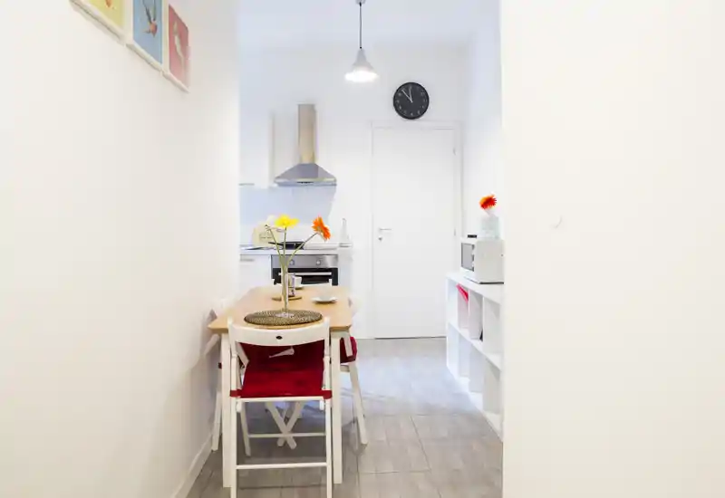 Zona pranzo luminosa e stretta con tavolo in legno per due, sedie bianche con cuscini rossi e fiori decorativi; angolo cucina moderno sullo sfondo.