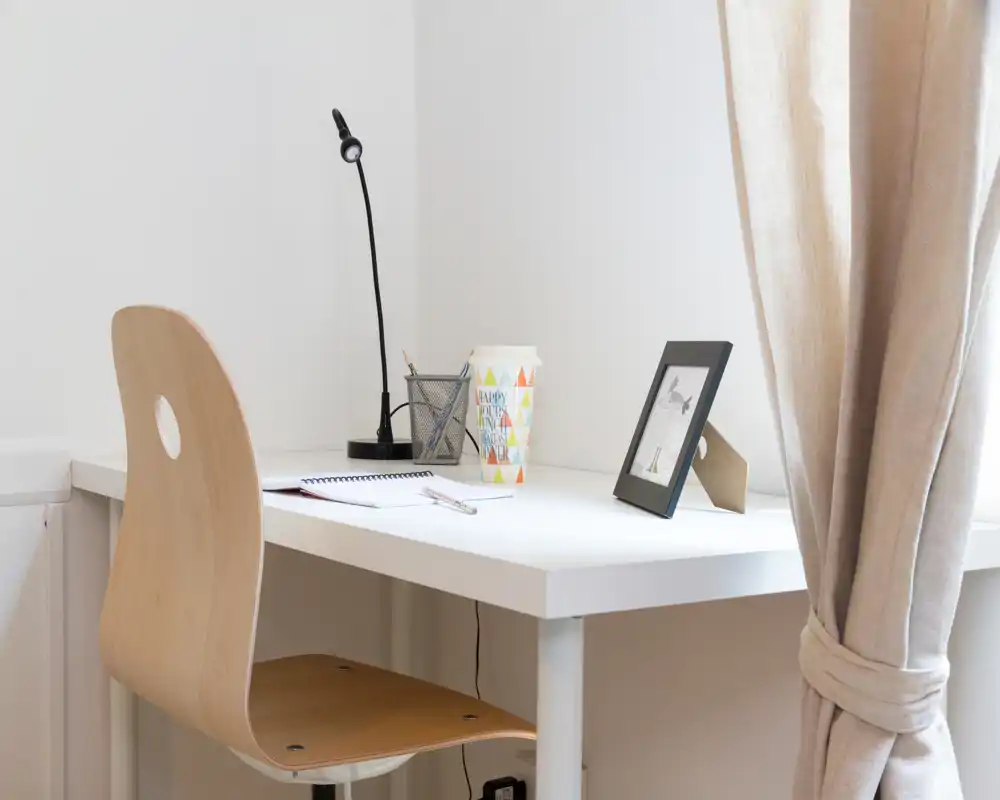 Angolo scrivania minimale e luminoso con sedia in legno, lampada da tavolo, quaderno e cornice: un'area lavoro ordinata vicino alla finestra con tenda.