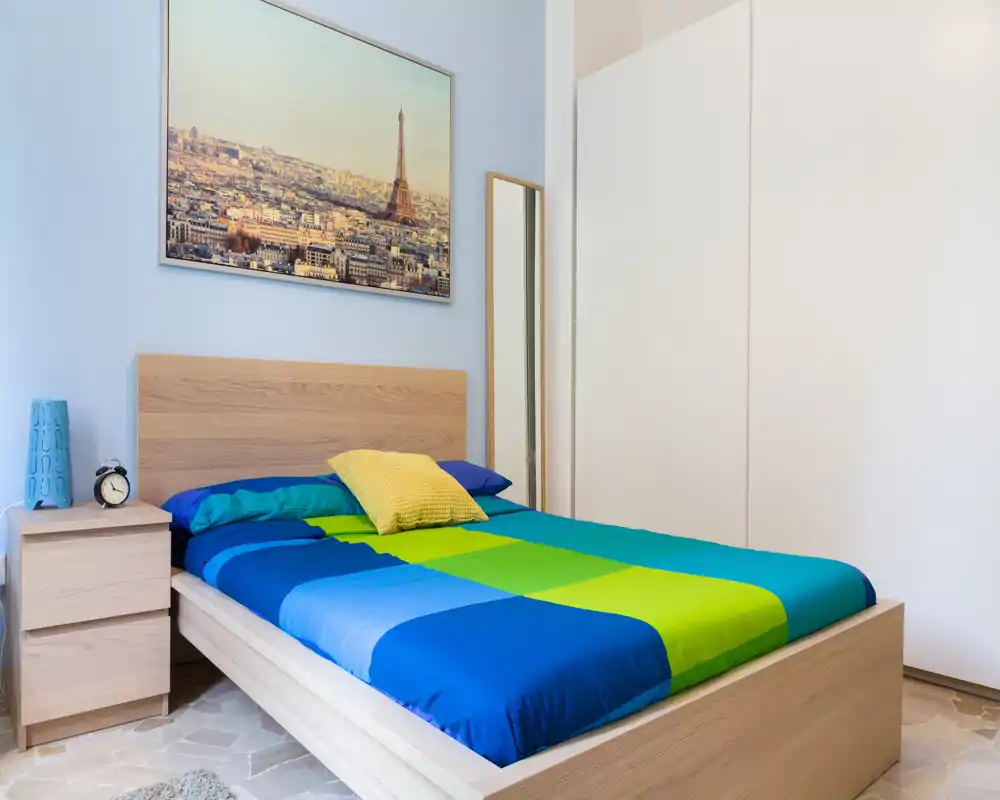 Camera da letto luminosa e ben allestita con struttura del letto in legno, biancheria colorata blu-verde e comodino con lampada e sveglia. Ampio armadio e quadro con stampa di Parigi completano lo spazio ordinato e accogliente.