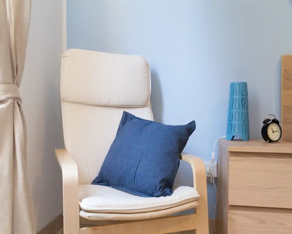 Angolo con poltrona chiara, cuscino blu e comodino in legno — un punto tranquillo ideale per leggere o rilassarsi.