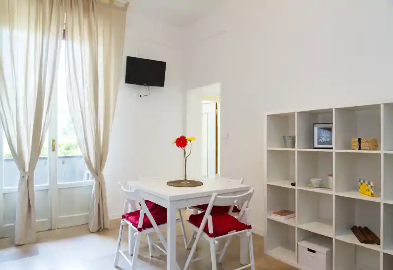 Bright, simple dining area with a white table, four chairs with red cushions and a decorative vase; shelving unit adds storage and styling next to large window doors.