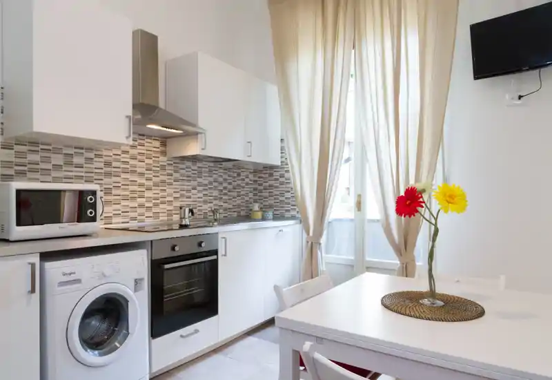 Bright, modern kitchenette with white cabinets, integrated oven and washing machine, a small dining table and natural light from a balcony door — ideal for a compact city apartment.