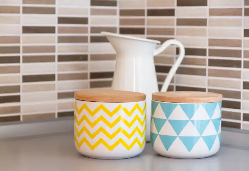 Close-up of a tidy kitchen counter with two patterned storage jars with wooden lids and a white pitcher against a tiled backsplash.