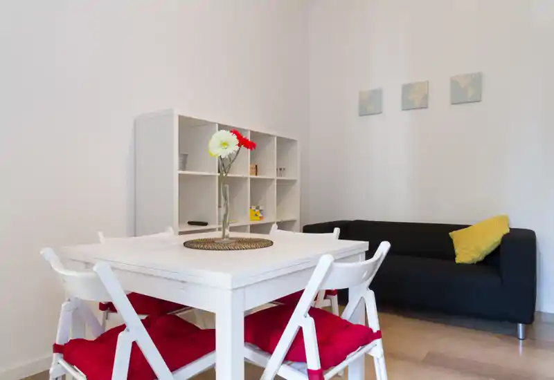 Well-staged dining area with a white table and four chairs dressed with red cushions, a decorative vase centerpiece and a comfortable black sofa in the background.