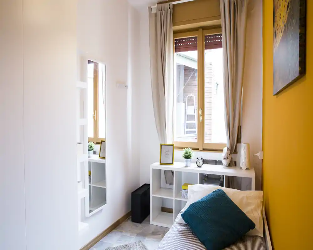 Sunny small bedroom with a single bed, teal cushion and warm yellow accent wall; a window with curtains and shelving under the sill create a practical, cozy corner.