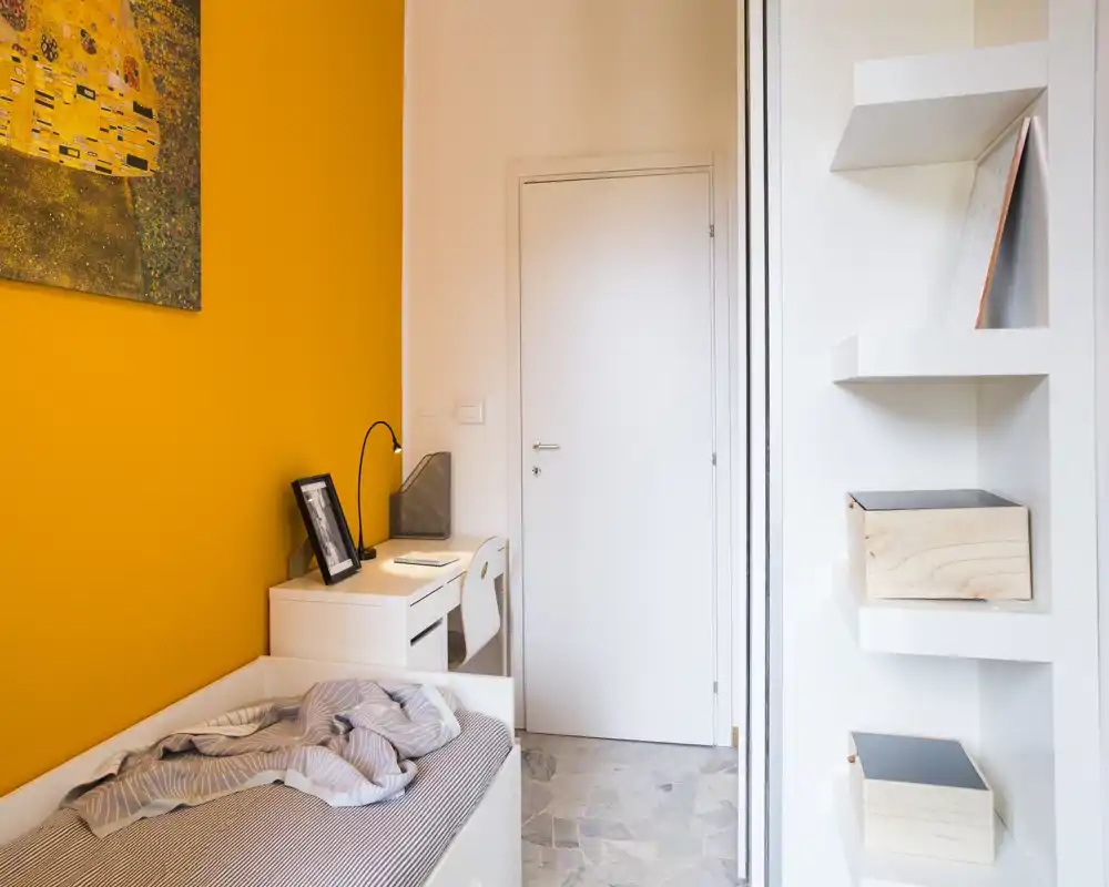 Sunny, modern small bedroom with a single bed against a bold orange accent wall, a compact white desk and shelving — bright and cozy setup ideal for a single occupant.