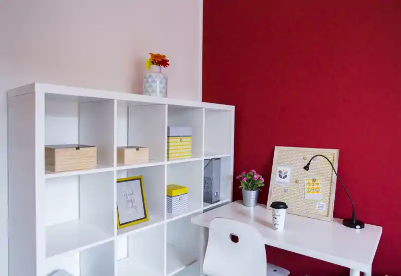 A tidy, styled workspace corner with white shelving, a compact desk and chair against a bold red accent wall; decorative boxes, a small plant and framed notes give a cozy, organized feel ideal for rental listing highlights.