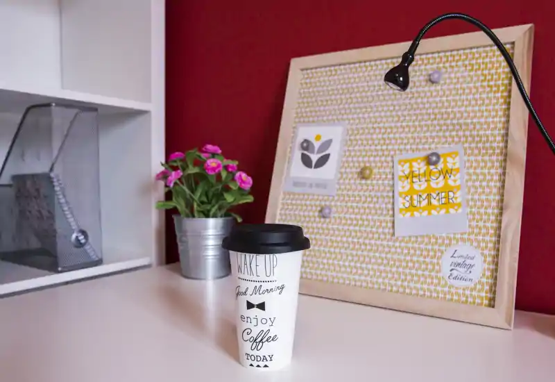 Bright bedroom corner styled as a small desk or vanity: a coffee cup on a white surface, decorative pinboard, pink flowers in a metal pot and a flexible lamp against a red wall.