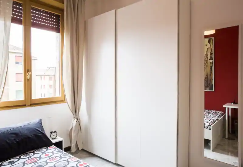 Chambre compacte et lumineuse avec linge de lit à motifs, table de chevet et grand placard. La lumière naturelle par la fenêtre et les rideaux neutres créent une atmosphère chaleureuse.