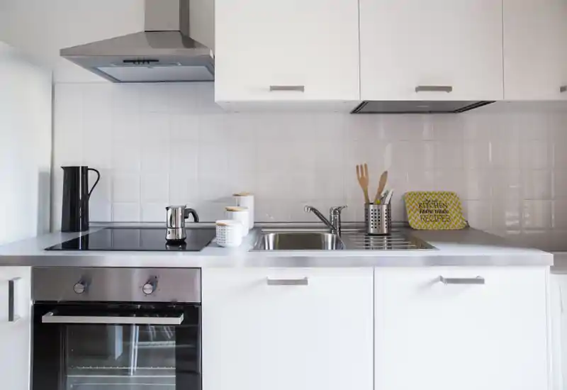 Bright, modern compact kitchen with an induction cooktop, built-in oven and stainless sink; minimalist white cabinetry and small decorative jars and utensils create a clean, welcoming cooking area.