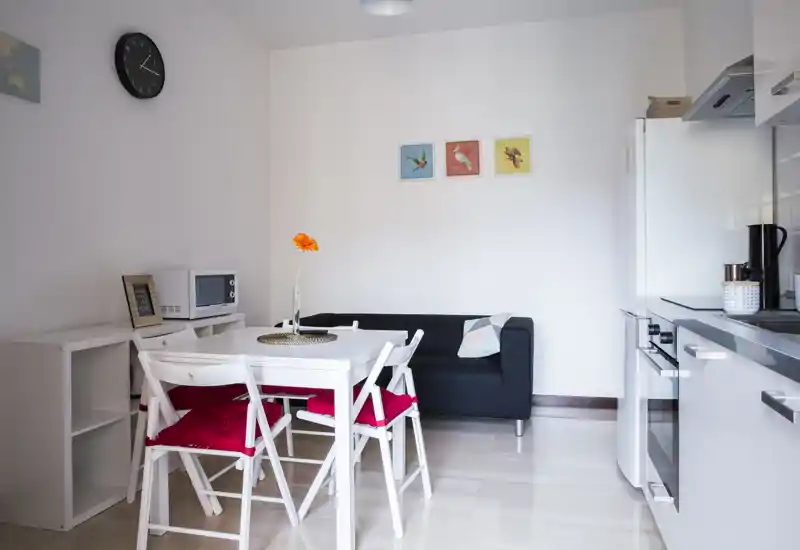 Bright, compact dining area integrated with a kitchenette: a small white table with red-cushioned chairs, a black sofa, and modern white kitchen units on the right. Minimalist décor with colorful wall prints and a single orange flower gives a fresh, welcoming feel.