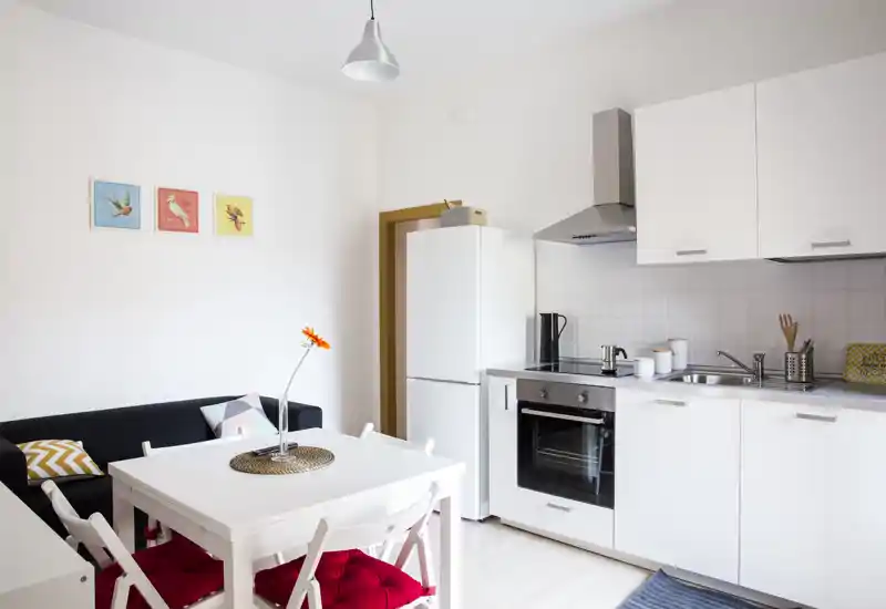 Bright, modern open-plan kitchen with white cabinets, built-in oven, fridge, sink and a small dining table with red-cushioned chairs — a clean, functional space ideal for short-term rentals.
