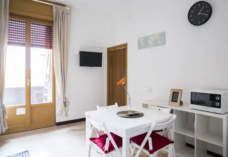 Bright, minimal dining area with a white table and four chairs, red cushions and a decorative centerpiece; large window/door brings abundant natural light.