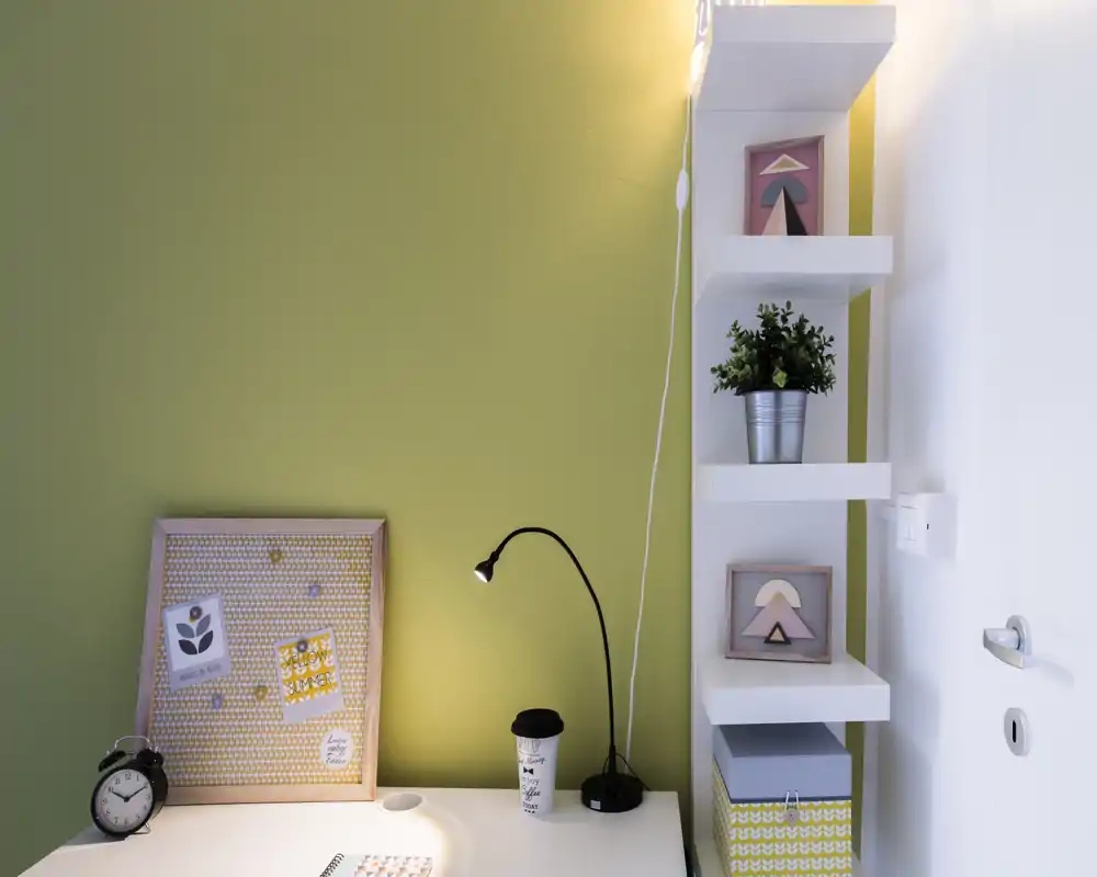 Angolo scrivania ordinato con lampada moderna, bacheca decorativa, pianta in vaso e mensole: un piccolo spazio di lavoro ben curato, ideale per studio o smart working.