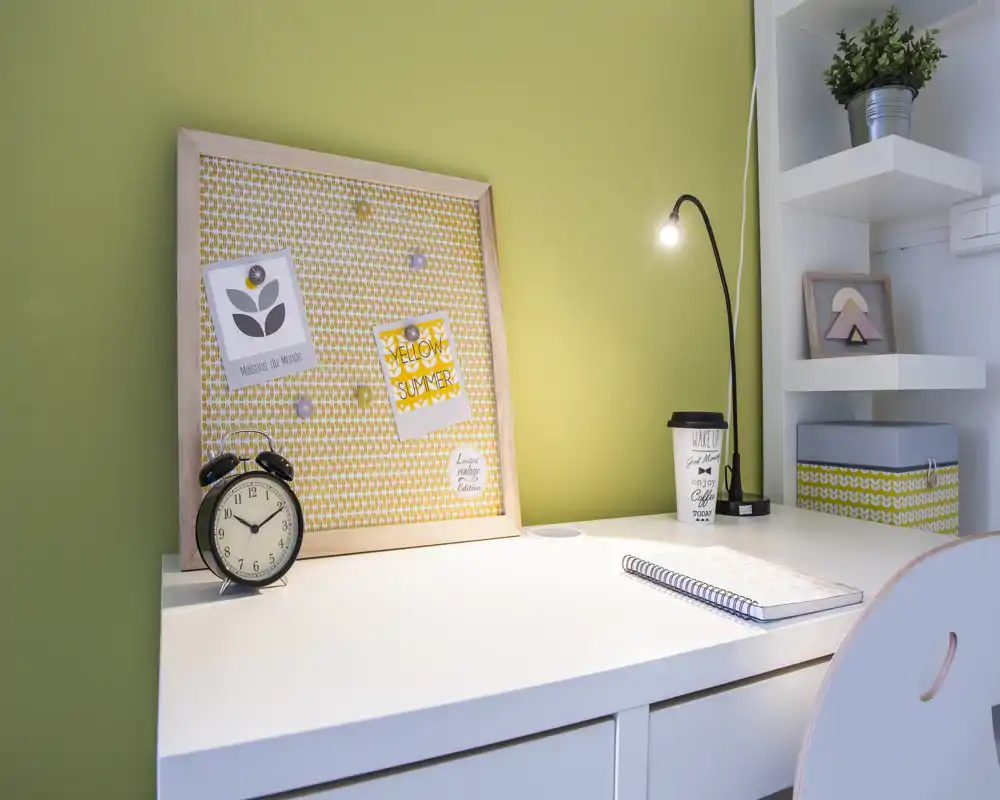 Angolo di lavoro luminoso e ordinato con scrivania bianca, lampada da tavolo, pannello memo con note, sveglia e scaffalatura; perfetto per studio o smart working.