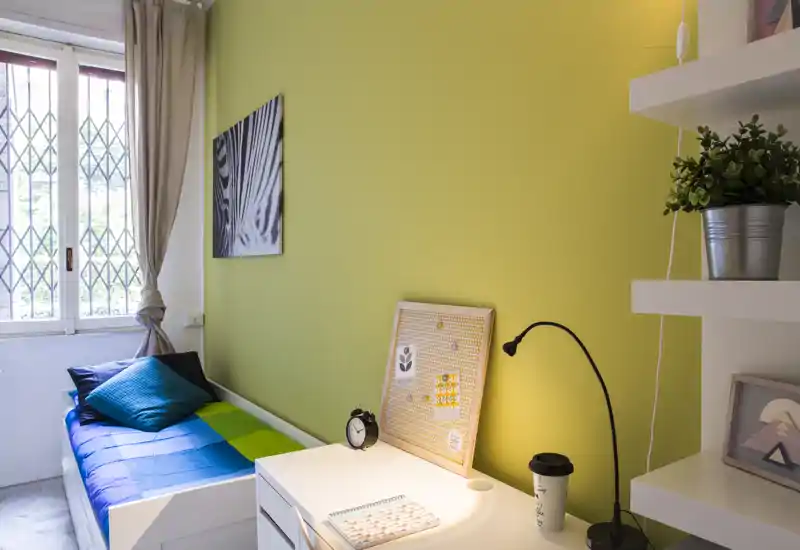 Camera singola luminosa con letto ordinato, scrivania con lampada da lavoro e mensole — ideale per uno studente o un occupante singolo.