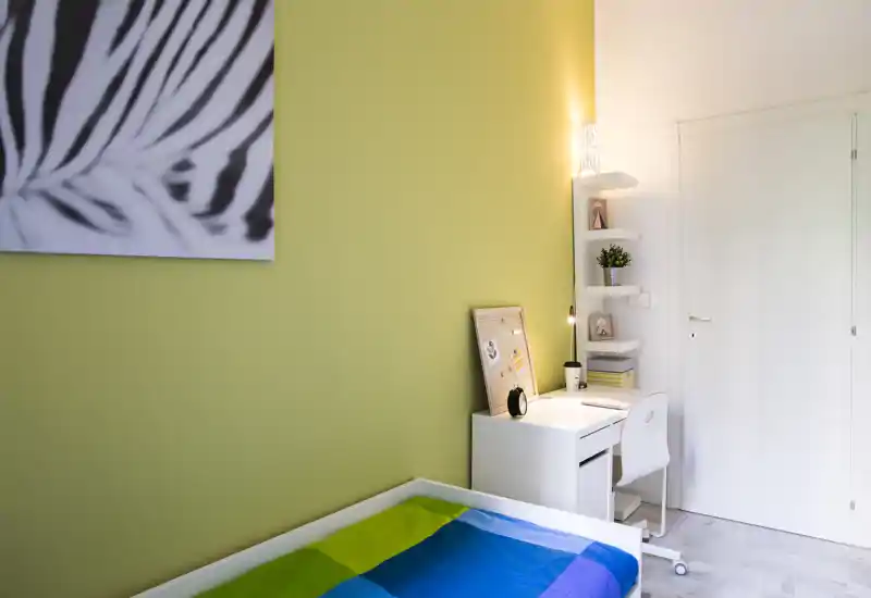 Camera singola luminosa e moderna con copriletto colorato, scrivania bianca compatta e mensole: ideale per uno studente o una persona sola.